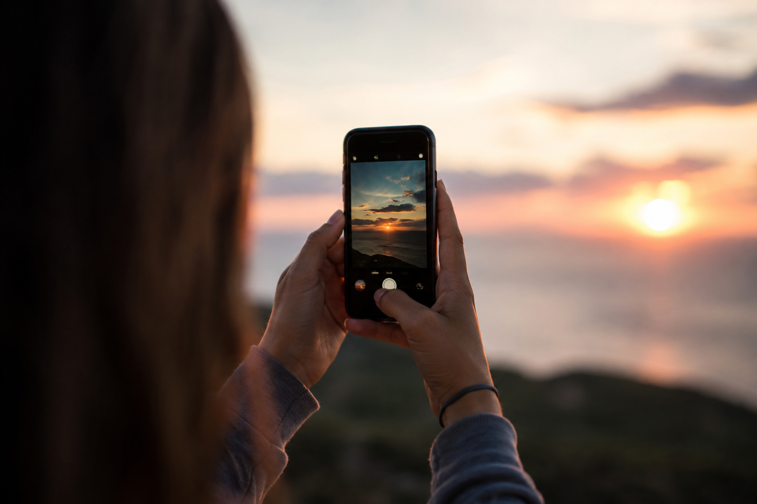 Persona sosteniendo un teléfono celular mientras fotografía un atardecer, con el paisaje desenfocado al fondo como símbolo de la relación entre la memoria y la tecnología.